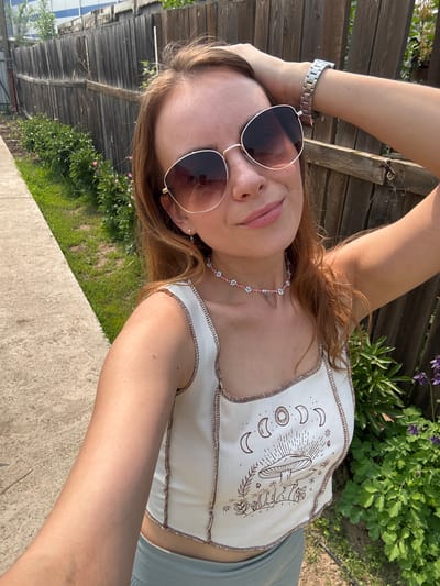 A young woman is taking a selfie outdoors in front of a wooden fence. She has long, reddish-brown hair and is wearing large, round sunglasses and a white crop top with a moon and mushroom graphic. She is also wearing a beaded necklace and a silver watch. The background shows a grassy area with some plants and flowers, and a concrete pathway. The overall lighting suggests it is a sunny day. The location is described as Tartat, Russia.