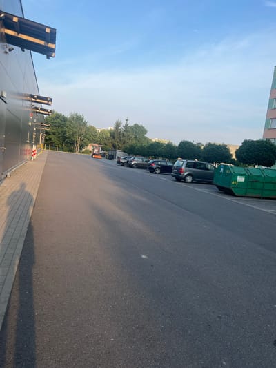 The image shows a parking lot adjacent to a modern building with a dark facade and horizontal lighting fixtures. Several cars are parked in designated spots, including a dark sedan, a black hatchback, and a dark gray station wagon. A large green dumpster with markings "013" is positioned on the right side of the parking lot. In the background, there are trees, a building with a multi-colored facade, and a clear sky with scattered clouds. A small red tractor or utility vehicle is visible further down the parking lot. The scene appears to be in an urban or commercial area.