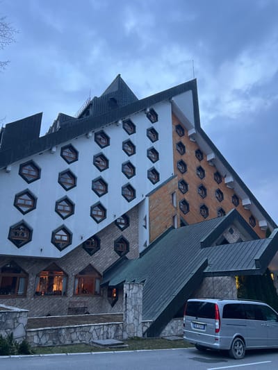 An exterior view depicts a large, angular building, identified as a hotel in Kolašin, Montenegro, under an overcast sky. The building exhibits a distinctive architectural style, featuring multiple stories with a complex, sloped roof structure clad in dark green material. The facade is sectioned, with a prominent white portion containing multiple rows of hexagonal windows with dark frames. An adjacent section features a brown wooden facade, also with hexagonal windows. A lower section is clad in light-colored stone, also incorporating hexagonal windows. The ground floor level displays larger, trapezoidal windows, with interior lights visible in some, casting a warm glow. A low stone wall with a flat top separates the building's immediate surroundings from a paved area in the foreground. Parked on this paved surface, facing away from the viewer towards the right, is a silver Mercedes-Benz van with the license plate "KE 919 KA". No individuals are visible. Bare tree branches are partially visible on the far left against the sky.