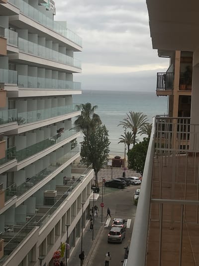 A high-angle, outdoor shot reveals a coastal cityscape in S'arenal, Spain. The view, likely from an elevated vantage point such as a hotel balcony, presents an overcast sky above a sea surface. Palm trees punctuate the line where the beach meets a road. Vehicles, including a police car with lights activated, are visible on the road alongside pedestrians. Modern architecture dominates the lower left quadrant, featuring a hotel with tiered, glass-fronted balconies. A contrasting older building, with white metal railings, is located in the upper-right. The scene implies a mix of tourism and local activity within an urban setting.