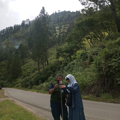 The image shows two people, a man and a woman, standing by a paved road that winds through a lush, green, hilly landscape. The man, wearing a dark vest over a blue shirt and dark pants, is holding a smartphone on a selfie stick and appears to be taking a photo. The woman, dressed in a light blue hijab and a striped blue dress, is looking at the phone. They are positioned on the side of the road, with a dense forest of trees and vegetation covering the hills behind them. The sky is overcast. The scene suggests a travel or sightseeing moment in a natural, outdoor setting. The location is identified as Kalalengkio, Indonesia.
