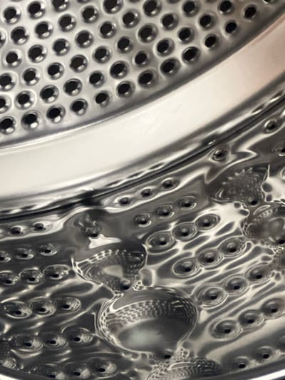 This is an extreme close-up, abstract image of the interior of a washing machine drum. The metallic surface is highly reflective, showing a pattern of circular indentations, or dimples, each with a small hole in the center. The lighting creates highlights and shadows that emphasize the curved and textured nature of the drum. The image is dominated by shades of silver and gray, with some areas of deeper shadow creating a sense of depth. There is a smooth, curved band running across the top of the frame, also reflecting light. The overall impression is one of industrial design and metallic texture. The image does not contain any information about Vanadzor, Armenia.