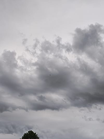 The image shows a cloudy sky above a treeline. The clouds are dark and ominous, suggesting that a storm may be brewing. There is a small object, possibly a bird or a drone, visible in the sky. The time of day is difficult to determine, but the overcast conditions suggest it may be later in the day or that the weather is poor. The location is identified as Kutná Hora, Czechia, but there are no specific visual cues in the image to confirm this. The overall mood of the image is somber and perhaps a little unsettling due to the dark clouds. There is no visible text in the image.