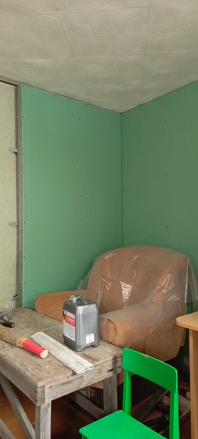 The image shows an indoor scene of a room under renovation. The walls are covered with green drywall, and the ceiling has a textured white finish. In the corner of the room, there is a sofa covered with a transparent plastic sheet. On a rustic wooden table in the foreground, a gray plastic container labeled "TIEFGRIJNT" is visible, along with some tools and building materials. A bright green chair is positioned next to the table. The overall impression is of an unfinished interior space. The image does not provide specific information about the city of Monastirische, Russia.