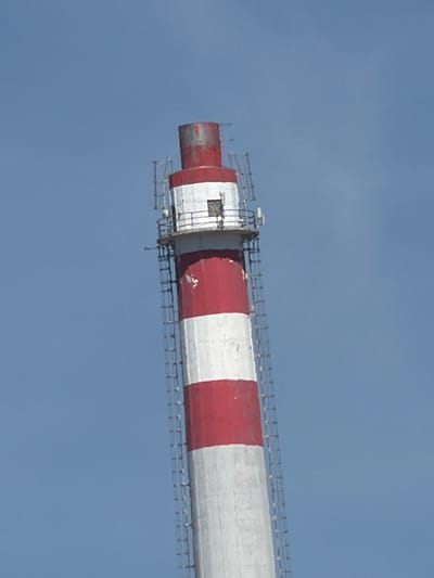 The image shows a close-up, side view of a large, cylindrical industrial smokestack against a clear, pale blue sky. The smokestack is painted with alternating red and white stripes. The stripes are horizontal and appear to be of equal width. The smokestack is constructed from metal.

Near the left end of the smokestack, there is a wider, red-banded section. This section appears to be an opening or a connection point, with some metal scaffolding or a platform attached to its underside. The scaffolding has a railing and appears to be made of metal struts. There are some rectangular white structures within this section, possibly indicating windows or access panels.

Further along the smokestack, the red and white stripes continue. The surface of the smokestack shows some signs of wear and tear, with areas of rust and chipped paint, particularly on the red sections. A thin metal railing or structure runs along the entire visible length of the smokestack, attached to its outer surface.

The lighting suggests it is daytime, with no shadows indicating the time of day or specific direction of sunlight. The sky is uniformly clear, suggesting fair weather. The context indicates the location is Olomouc, Czechia. There is no visible text in the image. The image focuses solely on the structure of the smokestack and its immediate surroundings against the sky, with no people or other objects present.