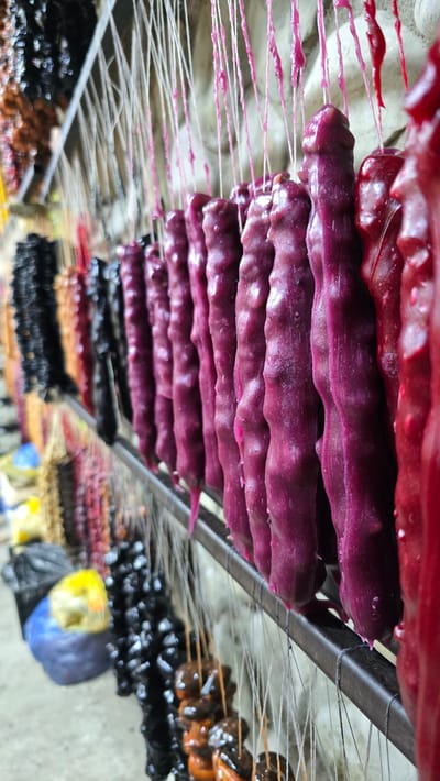 In Manavi, Georgia, a close-up shot captures a row of churchkhela, a traditional Georgian candy, hanging to dry. The candy consists of nuts, typically walnuts or hazelnuts, coated in thickened grape juice or fruit puree. The churchkhela are suspended by thin white strings from a metal rack, creating a visually striking display. The candies themselves vary in color, with prominent shades of deep purple and bright red, suggesting different fruit flavors or stages of drying. The texture appears slightly glossy and uneven, indicative of the coating process. In the background, out of focus, are other rows of churchkhela in different colors, such as black and a more muted brown or orange. The setting appears to be an outdoor market or a food production area, with a rough concrete floor visible at the bottom and a textured, light-colored wall or ceiling in the upper background. There are no people visible in the frame. The focus is sharply on the churchkhela, highlighting their form, color, and texture.