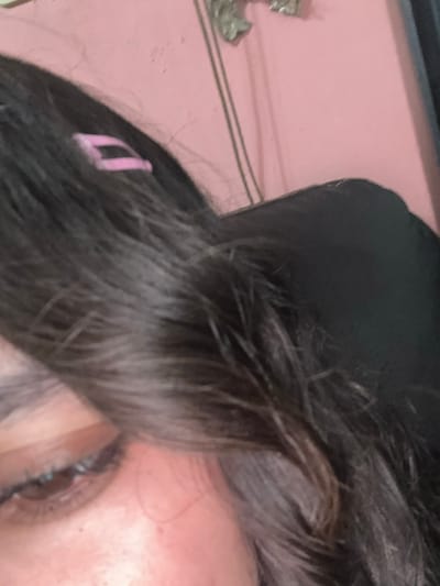 A close-up photograph shows a portion of a person's face. The person has dark brown hair with a pink bobby pin visible. The eye is visible, with dark lashes. The surrounding skin is fair. A black head covering is visible on top of the head. The background is pink. There are no discernible indicators of the location being San Cristóbal, Mexico.
