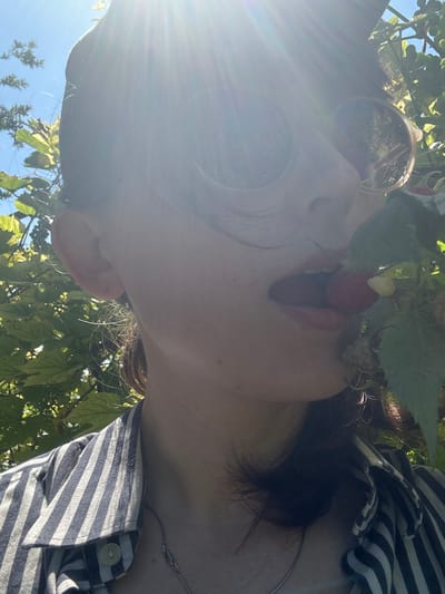 The image shows a close-up, low-angle shot of a person outdoors, surrounded by foliage and under a clear blue sky. The person is wearing round, reflective sunglasses and a striped shirt with a button. A raspberry is being held to their lips, suggesting they are eating or about to eat it. Sunlight streams into the frame, creating lens flares and highlighting the leaves and the person's face. The overall impression is one of enjoying a summer day and fresh fruit. The context provided indicates the location is Zvonarevka, Russia, and given the bright sunlight and lush greenery, it's likely daytime during a warm season.