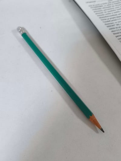 A sharpened green pencil lies on a white surface, angled from the top left to the bottom right. The pencil has a silver ferrule at its eraser end. To the right of the pencil, a book with blurred text is visible. The overall scene is neutral and appears to be an indoor setting, potentially a desk or table. There is no indication of the specific location in Rezina, Moldova, or any people or actions.