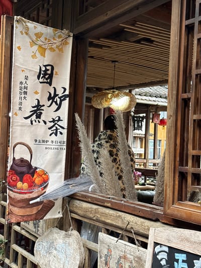 This image provides a detailed view of a traditional-style establishment in Gongtan, China, observed from an exterior perspective looking inward. The scene is characterized by dark brown, weathered wooden architecture, including a prominent window frame and a lower wooden railing. On the far left, a light-colored fabric banner hangs vertically from a wooden frame. It features yellow ginkgo leaf designs at the top and displays large black Chinese characters reading "围炉煮茶" (Boiling tea around a stove), accompanied by smaller Chinese text and the English phrase "BOILING TEA." Below this text, an illustration depicts a dark teapot on a grill, surrounded by persimmons and oranges in a wooden bucket.

Below the main window, a folded, grey umbrella rests on a wooden ledge, positioned above several rustic wooden signs with faded script and designs, which are suspended by twine. Through the open wooden window, the interior of the establishment is visible. A person, viewed from behind, stands or sits within the room, facing away from the viewer. They are dressed in a garment with a dark floral or patterned design on a lighter background. Above the person, an illuminated, oval-shaped pendant light fixture, constructed from woven material, hangs from a ceiling composed of wooden slats. In the foreground of the interior view, several tall, feathery, dried ornamental grass stalks are arranged, partially obscuring the lower part of the room. Further into the interior, additional traditional wooden structures, a red lantern, and parts of tiled rooftops from adjacent buildings are discernible in the background.