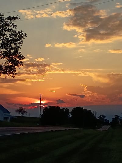 The image depicts a rural sunset scene in Lancaster, United States of America. The sky is ablaze with vibrant orange and yellow hues, with scattered clouds tinged by the setting sun. In the foreground, a dark silhouette of a tree branches out from the left side of the frame, partially obscuring the sky. A power line stretches horizontally across the upper portion of the image.

Below the sky, the landscape is characterized by a darkening environment. A white house with a dark roof is visible on the left, situated on a gentle slope. A lone tree stands on the hill to the right of the house. A utility pole with wires stands prominently in the middle-right of the frame, near the horizon. A paved road curves through the scene, leading into the distance. Dark green grass covers the lower portion of the image, with visible rows suggesting it has been recently mowed. The overall impression is one of a tranquil, rural evening.
