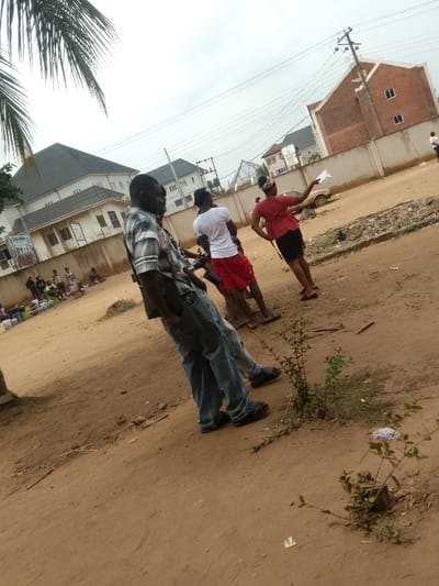 The scene depicts an outdoor urban environment in Umuokpu, Nigeria, characterized by an unpaved, reddish-brown dirt ground under an overcast sky. In the mid-foreground, a dark-skinned male subject is positioned, wearing a light-colored plaid short-sleeve shirt, blue jeans, and dark sandals. His left hand is in his front pocket, and his body is angled slightly to the right. Directly behind him and to his right, another male subject, partially obscured, is visible wearing a white t-shirt and red shorts, standing next to a dark-colored motorcycle. To the right of these two, a dark-skinned female subject, dressed in a reddish-orange short-sleeve top and a dark skirt, stands facing right. She holds a white sheet of paper in her raised left hand, gesturing or pointing to the right, with her right hand placed on her hip.
In the background, a concrete wall separates the dirt area from several buildings. To the left, a large multi-story building with a dark grey pitched roof and light-colored walls is visible. Further right, another similar building appears. On the far right, an unfinished multi-story building with exposed red brick walls and multiple rectangular windows is present. Utility poles with overhead power lines traverse the upper portion of the frame. A light-colored, older model car is partially visible behind the gesturing woman and the utility pole. To the far left, along the concrete wall, a group of approximately five to seven individuals are gathered, some seated on the ground near various containers and items, indicative of a market or street vending activity. Palm fronds extend into the upper left corner of the image, and small green shrubs grow from the dirt in the immediate foreground.
