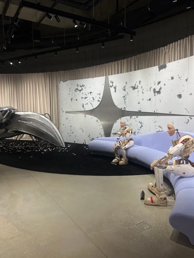 The image depicts an indoor exhibition space located in 성동구, South Korea. The foreground features a light gray concrete floor, which transitions to a large, circular black rug occupying the central and left portions of the scene.
On the left side of the black rug, a substantial, highly polished silver-metallic sculpture is positioned. This abstract form resembles a stylized bird's head, beak, or a futuristic vehicle's front, resting on thin support legs. Beneath this sculpture, numerous small, white or silver circular discs are scattered across the black rug.
To the right, two humanoid mannequins are seated on a curved, light blue upholstered sofa. These mannequins exhibit an unfinished or deconstructed appearance, with visible internal skeletal structures made of wood and metal strapping, particularly in their limbs and torsos. Their heads feature smooth, pale, simplified facial sections. Both mannequins wear tan-colored strapped footwear, resembling sandals or sneakers. One mannequin, seated closer to the foreground, shows a distinct reddish mark or stain on its lower right leg.
Behind the sofa and sculpture, a modern wall installation consists of large, light-colored panels with a textured, mottled gray pattern. A prominent, reflective metallic starburst-shaped element is integrated into the center of these panels. Above these panels, light beige or off-white curtains are drawn, extending across the upper part of the wall.
The ceiling is dark and exposed, featuring black overhead track lighting fixtures with multiple spotlights directed downwards. Visible ventilation pipes are also present on the ceiling. The overall lighting is artificial, creating a well-lit yet somewhat subdued ambiance within the space. No human individuals are present in the scene.