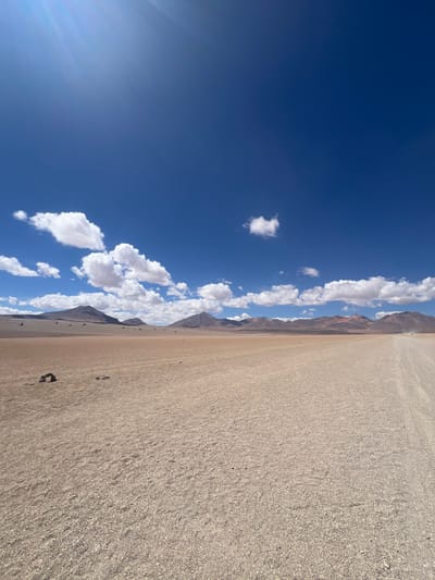 A desert landscape under a brilliant blue sky dotted with fluffy white clouds. The flat, sandy ground stretches to the horizon, where a series of low, brown mountains meet the sky. The sun shines brightly, casting a soft glow over the scene. A few scattered dark objects are visible on the desert floor.