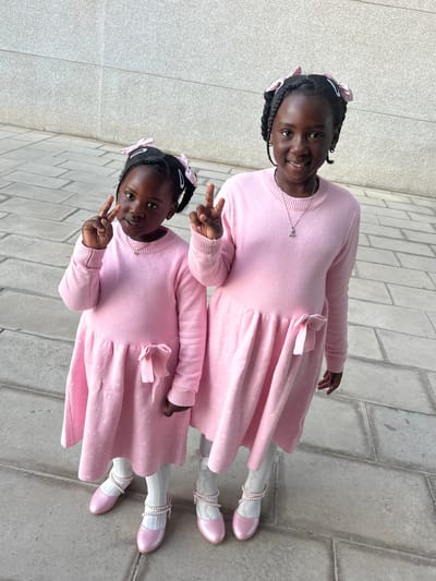 Two young girls, likely sisters or twins, stand on a paved surface, posing for a photo. Both girls wear matching light pink dresses with a subtle dotted pattern, long sleeves, and a decorative bow on the side. They also wear white tights and pink shoes adorned with pearl-like embellishments around the ankles. Both girls have their hair styled in braids with light pink ribbons. The girl in the foreground makes a peace sign with her right hand. The background consists of grey pavement, possibly a sidewalk, with subtle color variations that could indicate different paving stones. The location is potentially in Sant Vicent del Raspeig, Spain.