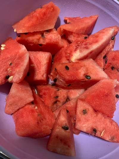 A close-up, overhead shot displays a purple bowl filled with chopped watermelon pieces. The watermelon is bright red and some pieces contain black seeds. The bowl is set against a blurred background that also appears to be purple. The scene is indoors. There is no indication of the location in Chiatura, Georgia.