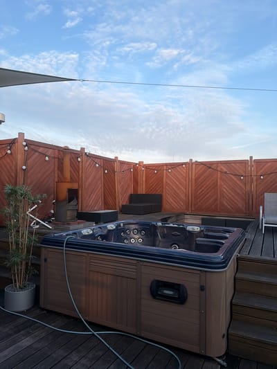 A wide shot captures an outdoor hot tub on a wooden deck, likely a rooftop or elevated patio, under a partly cloudy blue sky. The hot tub, a dark, multi-seat unit with visible jets, is positioned in the foreground. To the left of the hot tub, a tall, thin plant in a grey pot stands, with some white piping and hoses visible behind it. A corrugated metal outdoor heater or chimney unit is situated behind the plant and to the left of a wooden fence. The fence, made of dark wood with a chevron pattern, encircles the deck and is adorned with a string of white globe lights. In the background, dark outdoor furniture, including a sectional sofa, is visible. A white lounge chair is also present on the right side of the deck. The overall setting suggests a private outdoor relaxation area. The scene is devoid of people.