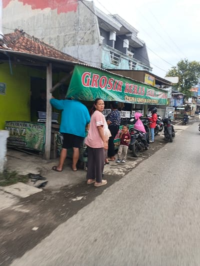 The image depicts a street scene in Cangkuang Kulon, Indonesia, outside a rice wholesale establishment. On the left, a single-story building with a lime-green facade and a brown tiled roof features a prominent green banner with red lettering reading "GROSIR BERAS KILLA" (Wholesale Rice KILLA) and "TERIMA GROSIR DAN ECERAN" (Accepts Wholesale and Retail). Below it, another green banner shows "JL PUTRA DUA PUTRI NITA'Z" and a phone number. Price signs are visible beneath the main banner.

Approximately seven individuals are present. In the mid-ground, a woman wearing a light pink t-shirt and dark pants, holding a light-colored bag, stands facing the camera. Behind her, a person in a blue long-sleeved shirt and black shorts, wearing sandals, stands with their back to the viewer, facing into the shop. Another woman in a dark patterned top is partially visible behind the first woman. Next to her, a small child in a maroon t-shirt and light-colored pants consumes an item, looking towards the right. Further right, a person in a red jacket and helmet stands near a parked black motorcycle that has a pink wrapped package secured to it. More motorcycles are parked along the curb, extending down the street.

The street comprises a paved asphalt road in the foreground and a concrete sidewalk/curb separating it from the buildings. Beyond the shop, a taller, multi-story building with a light blue/grey facade and white decorative architectural elements is visible. The sky is overcast. Signs of wear are present on the buildings. Debris and irregular ground are visible on the sidewalk in the immediate foreground.