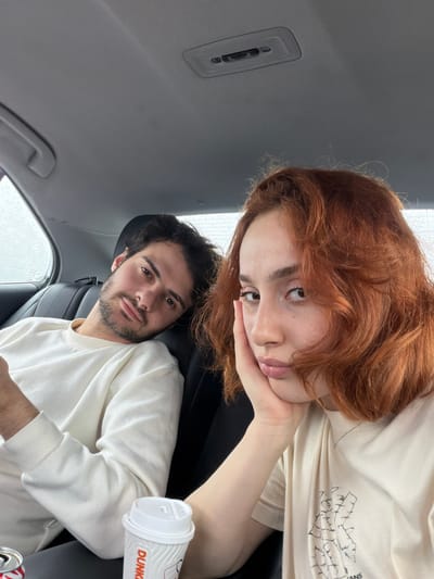 The image depicts two people in a car, likely parked. A woman with red hair is positioned in the upper part of the frame, resting her face on her hand. She is wearing a light-colored t-shirt. A man with dark hair and a beard is in the lower part of the frame, also wearing a light-colored sweater. He is looking towards the camera. A Dunkin' coffee cup is visible near the man. Raindrops are visible on the car windows. The interior of the car appears to be modern, with gray upholstery and black leather seats. The specific location within the city of Ianeti, Georgia cannot be determined from the image.