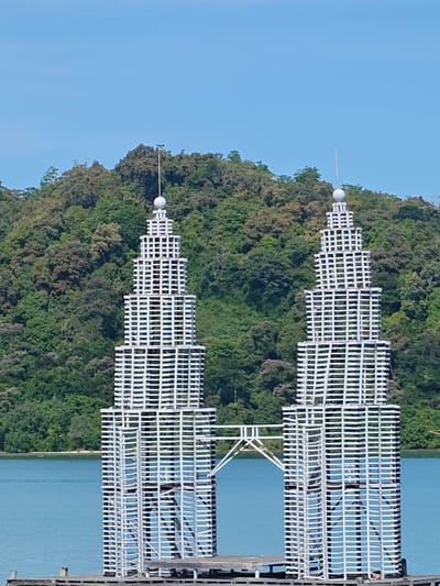 The image depicts a scaled-down model of the Petronas Twin Towers, constructed from white latticework, situated on a platform in a body of water. A bridge-like structure connects the towers. Lush green foliage covers a hillside in the background. The sky is clear and blue. The towers are identified as being located in Layeun, Indonesia.