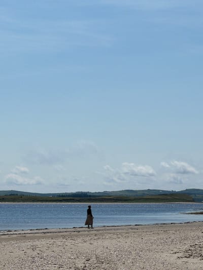 The image depicts a solitary figure standing on a sandy beach. The person is facing away from the camera, looking out towards a body of water. The figure is wearing a dark top and a long, light-colored skirt or dress. The beach is composed of sand and small shells. Beyond the water, a green, hilly landscape stretches across the horizon, with some scattered trees and buildings visible. The sky is a clear, pale blue with a few wispy clouds. The scene appears to be a coastal area in Tullycullion, Ireland, under a bright, daytime sky.