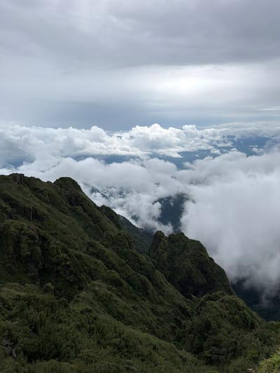A wide-angle, high-angle shot captures a majestic mountain landscape in Bản Bo, Vietnam, under a cloudy sky. The foreground is dominated by the rugged, dark green slopes of a mountain range, covered in dense vegetation and appearing mossy in places. Sharp peaks and deep ravines characterize the terrain. In the midground and background, thick, white clouds create a surreal effect, obscuring the lower parts of the mountains and forming a sea of clouds. Beyond the clouds, hints of more mountain peaks and valleys are visible. The sky is overcast with layers of gray and white clouds, with a brighter area suggesting sunlight filtering through in the distance. The scene is devoid of people or any man-made objects.
