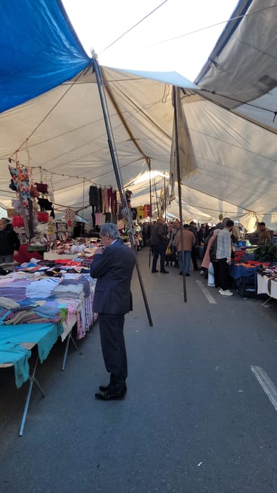 An outdoor marketplace in Beylikdüzü, Turkey, shows a man in a dark suit and black shoes examining merchandise on a table, his head bent low. The table is piled with clothes and textiles in a variety of colors. Above, a large white tent provides cover for the market, and the open sky is visible to the upper left. Other people browse the market, and additional tables of goods are arranged along the thoroughfare. The ground is paved.