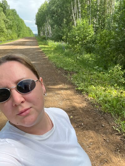 A selfie is taken by a woman with blonde hair and sunglasses, wearing a white t-shirt. She is standing on a dirt road lined with trees on both sides. The road appears to be in a rural or natural setting. The sky is visible and appears partly cloudy. The woman is positioned in the foreground, with the road and trees extending into the background. The scene is illuminated by natural sunlight.