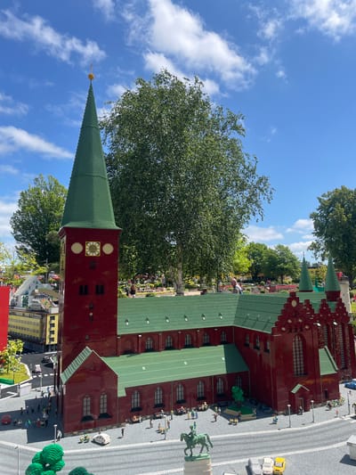 This is a high-angle, close-up, eye-level, medium shot of a Lego model of a brick church with green roofs, located in Billund, Denmark. The church has a tall, slender tower with a clock and a pointed green spire topped with a gold orb. The main body of the church has a pitched green roof and numerous arched windows. The surrounding area is also made of Lego, featuring miniature buildings, trees, cars, and people. The sky above is blue with scattered white clouds, and some real trees are visible in the background. The scene depicts a sunny day in what appears to be a miniature park or attraction.