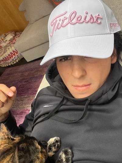 The image depicts a close-up, chest-up view of an individual in an indoor setting, identified as Fraccionamiento y Club de Golf los Encinos, Mexico.

**People:** One individual is present, a young adult with dark hair, looking directly at the camera with a neutral expression. They are wearing a white Titleist Pro baseball cap with pink embroidered lettering, and a black Nike hooded sweatshirt featuring a white swoosh logo on the left chest. The black drawstring for the hood is visible. The person's left hand, with trimmed fingernails, is partially visible in the lower-left corner.

**Subjects/Objects:** A small animal, identified as a cat with a tortoiseshell coat pattern (patches of brown, black, and tan/cream), is resting on the individual's chest/lap in the lower-left portion of the frame. Two of its paws, exhibiting similar mixed coloring, are visible on the black sweatshirt.

**Environment:** The background suggests a domestic interior. A light-colored sofa or armchair with a patterned cushion (featuring shades of orange, red, and cream) is visible behind the individual. A multi-tonal rug or carpet, primarily purple and brown, is visible on the floor. A light-colored wooden surface (possibly flooring or wall paneling) is present on the far left.

**Actions/Interactions:** The individual appears to be in a relaxed, reclined position, likely taking a selfie or being photographed. The cat is resting on the individual, indicating a passive interaction of proximity or comfort.