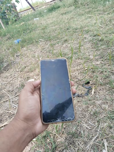 The image shows a hand holding a damaged smartphone outdoors. The phone's screen is cracked and reflective. The background consists of dry grass and some green vegetation. A discarded blue plastic bottle is visible in the distance. The location is Gebang, Indonesia.