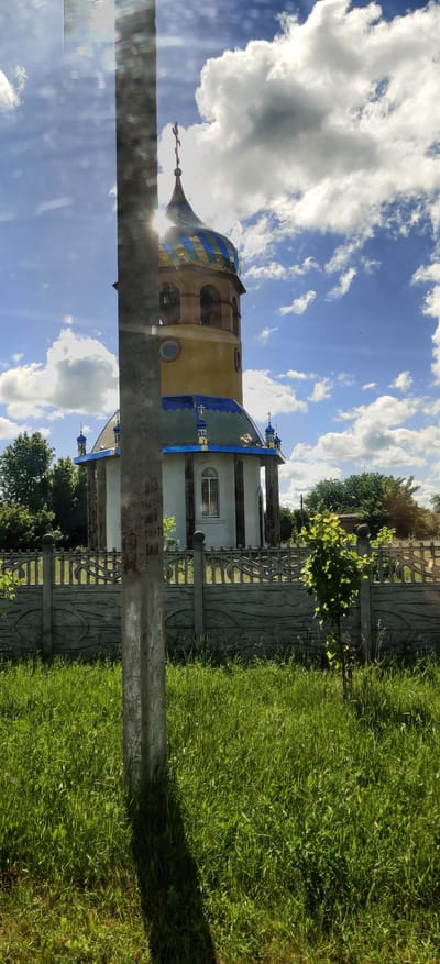 The image shows a church with a blue and yellow domed roof, located in the village of Shelestove, Ukraine. A concrete fence with an ornate pattern runs across the foreground, with tall green grass in front of it. A utility pole stands to the left of the frame, casting a shadow. The sky is blue with fluffy white clouds. The sun is shining brightly, creating lens flare in the upper left portion of the image.