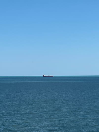 The image depicts a vast expanse of the Black Sea under a clear, light blue sky. In the distance, a solitary cargo ship with a red hull is visible, its form small against the immense horizon. The water is a deep blue, with subtle ripples indicating a calm surface. The scene appears to be a wide shot taken from the shore or a vantage point overlooking the sea, characteristic of the coastline near Kryzhanivka, Ukraine, a coastal settlement on the outskirts of Odesa. There are no people or immediate actions depicted, conveying a sense of isolation and the tranquil, expansive nature of the maritime environment.