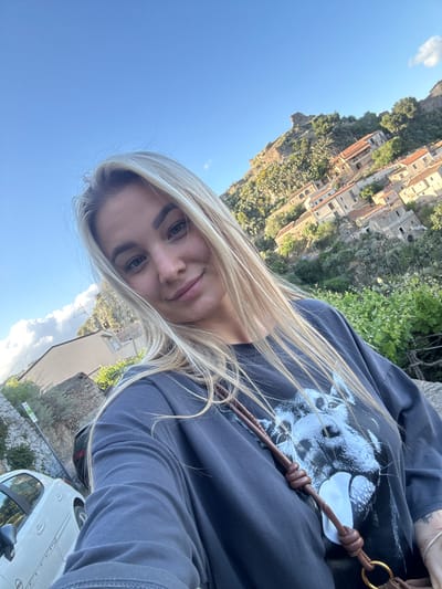 A young blonde woman wearing a dark sweatshirt with a graphic print is taking a selfie. The location is Rina, Italy; a cityscape of buildings on a cliff is visible in the background to the left. The sky is blue. A portion of a white car is in the lower right corner.
