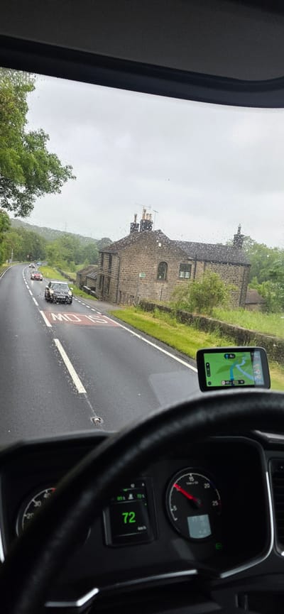 The image captures a view from inside a vehicle, looking through the windshield on a rainy day. The road ahead is wet, with visible raindrops on the glass. A stone-built house with a slate roof and chimneys stands prominently on the right side of the road. Trees and green hills form the background under an overcast sky. Several cars are visible on the road, including a black Mini Cooper directly in front of the vehicle. A "SLOW" road marking is painted on the asphalt. A GPS device is mounted on the dashboard, displaying a navigation map. The vehicle's dashboard shows a digital speedometer reading "72" and an analog tachometer. The overall scene suggests driving through a rural or semi-rural area in overcast weather. The location is identified as Barnsley, United Kingdom.