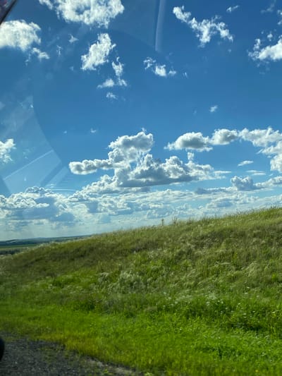 This is a photograph taken from inside a vehicle, looking out through the windshield. The scene depicts a bright, clear day with a vibrant blue sky filled with fluffy white cumulus clouds. The lower portion of the image is dominated by a lush green grassy hillside, possibly a rural landscape in the vicinity of Kaldarovo, Russia. In the distance, rolling hills and fields can be seen under the expansive sky. There are no people or discernable actions within the frame. The overall impression is one of natural beauty and open space.