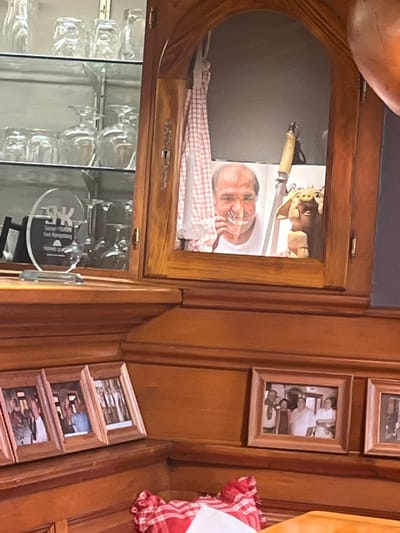 The image depicts the interior of the Restaurant-Bar Karl Ammelburg in Sélestat, France, identified by a discernible "Karl-Ammelburg Trophée Qualité" plaque. The environment is characterized by polished wooden cabinetry and paneling.

On the left, a glass-fronted display cabinet houses multiple clear drinking glasses, including tumblers, stemware, and decanters, arranged on glass shelves. A clear oval acrylic award plaque, featuring a "JK" logo and partial text "Suisse," "EUROPE," and "Karl-Ammelburg Trophée Qualité," is prominently displayed on a lower shelf.

Centrally, a wooden cabinet with an arched opening frames an image or reflection of a middle-aged male with a receding hairline, holding a clear glass to his mouth. Within this framed area, a red and white checkered cloth hangs on the left side, a knife with a light-colored handle is positioned vertically behind the man's head, and a small, light-brown animal figurine, resembling a pig, stands to the man's right.

Below these cabinets, a wooden ledge holds a series of small, light-wood framed photographs, each depicting groups of people. In the bottom right foreground, a section of a table is visible, covered by a red and white checkered cloth, with a piece of white paper resting upon it.