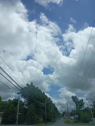 The image captures a view of a suburban street in Nicholasville, Kentucky, USA, under a dynamic sky filled with fluffy white clouds against a bright blue backdrop. The perspective is from inside a vehicle, looking forward and slightly upwards. Overhead power lines crisscross the frame, supported by utility poles that dot the sides of the road. Lush green trees line the street, their foliage dense and varied, with some appearing as large, mature trees and others as more conical evergreens.
On the left side of the road, a small section of a brick house with white trim is visible, suggesting residential properties. A street sign, partially obscured by the foliage and a utility pole, reads "HEATHCOTE". Further down the road, two vehicles are traveling away from the camera, one a dark-colored sedan and another a lighter-colored vehicle. Yellow traffic signs, including a warning sign indicating a curve or turn, are also present along the roadside. A bright red fire hydrant is visible near the base of some of the trees on the left. The road itself is paved and appears to be a two-lane street, gently curving and sloping slightly uphill. The overall impression is a typical, quiet suburban environment on a partly cloudy day.