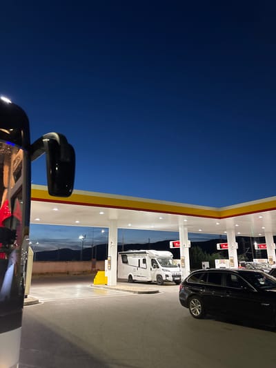 The image depicts a Shell gas station in Kalotina, Bulgaria at dusk or night. Several vehicles are parked at the pumps, including a black car and a white RV. The gas station's structure is clearly visible with its distinct Shell logo. The scene is illuminated by artificial lighting. A vehicle is partially visible in the foreground, with its side mirror prominent in the bottom left corner of the frame. The sky is a gradient of blue, indicating the time of day.