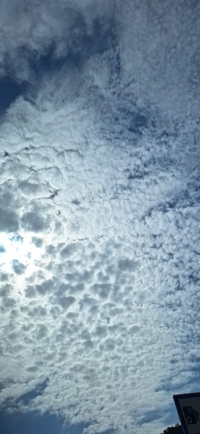 The image shows a sky filled with altocumulus clouds, with a patch of bright light likely from the sun shining through. A small portion of a signpost is visible in the top right corner. This image depicts a sky in Cherniche, Bulgaria.