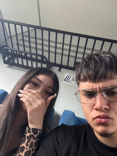 Two individuals are depicted in a close-up, likely a selfie, seated within a public transport vehicle in the city of Trastikovo, Bulgaria. The female on the left has long, dark hair and wears a leopard-print patterned sleeve. Her right hand, featuring light-colored manicured nails, is placed over her mouth, partially obscuring her face, while her left eye is visible looking towards the camera. The male on the right has short, dark hair with visible lighter streaks, a fringe haircut, and wears a black t-shirt. He wears eyeglasses with thin metal frames and has a small patch of hair below his lower lip. His expression is direct, with slightly pursed lips and a furrowed brow, looking towards the camera. Both are seated in blue, perforated upholstered seats. The background features a light-colored wall with a black metal railing containing vertical bars. A white rectangular sign with purple graphical elements, including two QR codes and partial Cyrillic text "ЧЕТИ ВР", is affixed to the wall behind the subjects.
