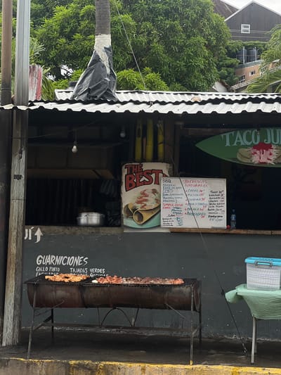 The image depicts a street-side food stand in San Juan del Sur, Nicaragua. The food stand features a corrugated metal roof with visible lighting fixtures. In front of the stand, a large grill is set up, loaded with various meats, possibly pork and chicken. To the left of the grill, text on the wall reads "GUARNICIONES GALLO PINTO TAJADAS VERDES ENCILADAS NACHOS". A menu board is also visible, listing items like "BBQ CHICKEN PORK OR BEEF", "REFRESCOS NATURALES", and various types of tacos and burgers, with prices indicated. A cooler filled with what appear to be stacked plates or containers sits on a table to the right of the grill. The background shows lush green trees and buildings, characteristic of the tropical environment of San Juan del Sur. The overall scene suggests a vibrant, open-air eatery preparing to serve customers.