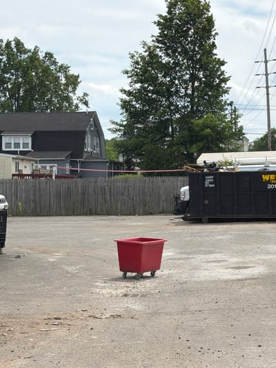The image depicts an outdoor scene in Norwood, United States of America, under a partially cloudy sky. In the foreground, a red wheeled bin is positioned in a gravelly area. To the left, a black dumpster is filled with debris, including wooden pallets and other materials. Behind the dumpster, a white truck is partially visible. A weathered wooden fence runs across the middle ground. Beyond the fence, a gray house with a dark roof is seen, with trees and utility poles in the background. The overall environment appears to be an urban or semi-urban setting, possibly a commercial or industrial lot. There are no people visible in the image.