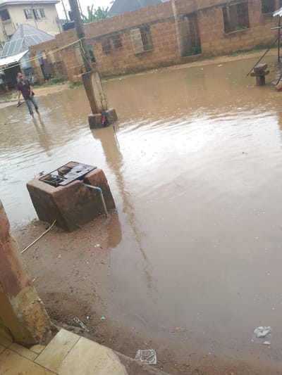 The image depicts a flooded street in Atughobi, Nigeria. A man in a red cap and blue jeans is walking through the brown, murky water, holding a pair of black shoes in his hand. He appears to be wading through waist-deep water. In the background, there are brick buildings with open windows and a partially constructed structure with solar panels on its roof. A utility pole stands prominently in the middle of the flooded area, with wires running across it. A concrete structure, possibly a well or a water pump base, is submerged in the water in the foreground. The water level is high, indicating significant rainfall. The ground surrounding the water is muddy and littered with debris, including plastic bags and sticks. The overall scene conveys the impact of flooding on the urban environment.