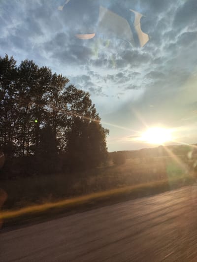 The image depicts a landscape scene with a road in the foreground, likely in or near the city of Diyashevo, Russia. The sun is setting, casting a bright glow and lens flares across the sky and the landscape. Trees are visible on the left side of the image, silhouetted against the bright sky. The sky is partly cloudy, with the sun prominently positioned on the right side of the frame, creating a starburst effect. The road surface shows motion blur, suggesting the photo was taken from a moving vehicle. Reflections are visible in the upper portion of the image, possibly from a window. The overall impression is of a scenic sunset viewed from a roadside.