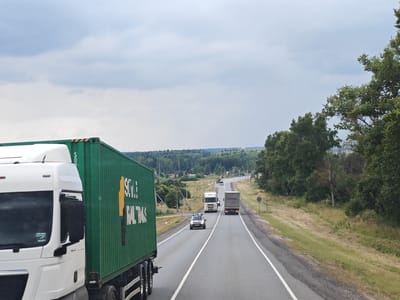 The image captures a highway scene in Kul'tura 1, Russia, under a cloudy sky. In the foreground on the left is the side of a white truck cab and a large green shipping container with yellow lettering that reads "SOYUZ TRANS". The highway extends into the distance, with several other vehicles including trucks and a white car. The roadside is bordered by lush green trees and vegetation, with a slightly undulating landscape. The overall impression is of a journey on a rural highway on an overcast day.