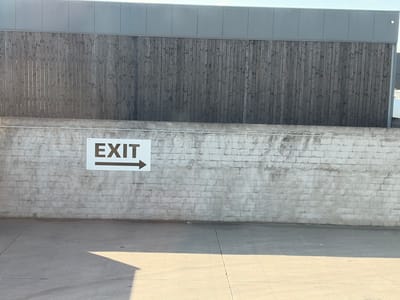 The image depicts an outdoor scene featuring a light-colored brick wall with an "EXIT" sign and an arrow pointing to the right, situated on the wall. Above the brick wall is a dark wooden fence, and atop the fence is a gray metal structure. The ground in front of the wall is paved with concrete, with a shadow cast on it from an unseen object. The scene appears to be located in the city of Veurne, Belgium, likely in an industrial or commercial area, possibly a parking lot or access road. No people are visible in the image.