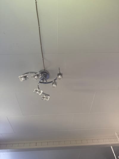 A modern chrome chandelier with five clear glass shades hangs from a white, textured ceiling. The chandelier is suspended by a thin wire that runs up towards the ceiling. The shades are cubic in shape with internal light fixtures. The ceiling is divided into rectangular panels by subtle seams. Below the ceiling, there is a decorative white trim or molding. The image is taken from a low angle looking upwards. The location is a room in Moelv, Norway.