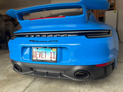 The image shows the rear of a bright blue Porsche 911 Carrera GTS parked in what appears to be a garage. The license plate reads "DE VET 11" and is from Illinois, with "LAND OF LINCOLN" printed below. The car has a prominent rear wing and dual exhaust pipes. In the background, to the left, part of a dark-colored SUV is visible. To the right, there is a white refrigerator and wooden shelving with items on it. The environment is a concrete floor and walls, suggesting an indoor setting like a garage. There are no people visible in the image.