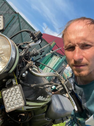 A man, appearing to be in his late 30s to early 40s, with thinning brown hair and a light beard, is taking a selfie in front of a motorcycle. He is wearing a teal t-shirt with a camouflage pattern. The motorcycle, a black Maverick, is partially visible, showing its headlight, handlebars, and a square LED auxiliary light. The background features a rustic wooden fence with a red-roofed building behind it, under a bright blue sky with wispy clouds. The scene appears to be outdoors, possibly in a rural or semi-rural setting in Nizhnii Bugrysh, Russia, given the context. The man's expression is neutral, and the overall impression is that of a casual, possibly impromptu, photograph.